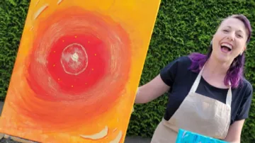 A woman presents a bright, artistic painting outdoors.
