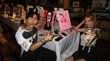 Boy points surprised at a painting, girl drinks cocktail.