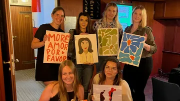 Six women show self-painted pictures in a room.