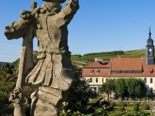 Steinskulptur steht, während Weinberge und Gebäude im Hintergrund sichtbar sind.