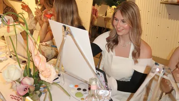 A woman is painting on a canvas with a smile, surrounded by friends.