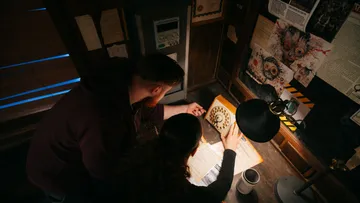 Zwei Personen untersuchen eine mysteriöse Maschine am Tisch.