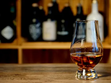 A whiskey glass stands on a wooden table with bottles in the background.