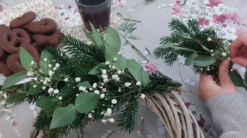 Woman creates a wreath with green branches and flowers.
