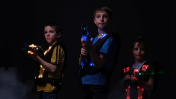 Three children hold glowing toy glass guns in the dark.