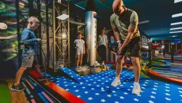 Man putts golf, children watch in colorful interior.
