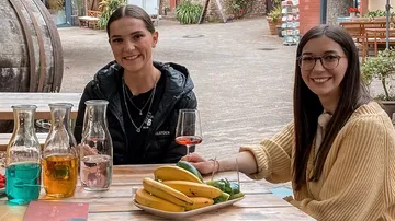 Two women sit at a table with drinks and bananas.
