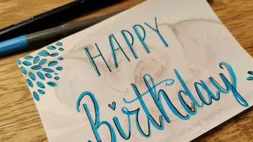 Handgemachte „Happy Birthday“ -Karte mit blauem Text und Blumenkritzeleien auf einem Holztisch.