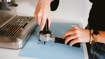Hands squeeze coffee powder in a portafilter on a work surface.