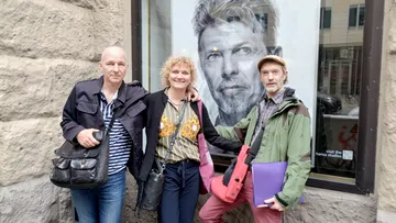Drei Personen stehen neben einem Porträt an einer Wand.