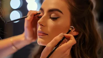 A woman receives makeup with brushes in a bright room.