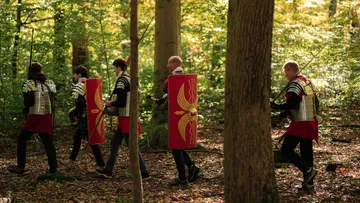 Four people in Roman armor walking through forest.