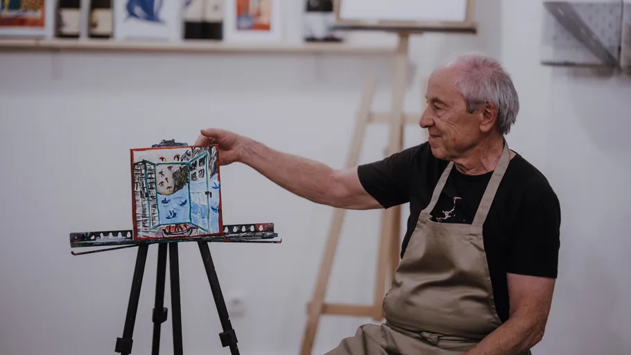 Elderly man in apron displays artwork on easel.
