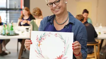 Frau, die ein Aquarellkranz in der Hand hält und lächelt.