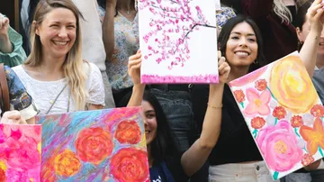 Women holding colorful floral paintings, outdoors.