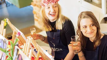 Two smiling women holding drinks at a painting class.