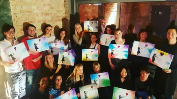 Group holding paintings of girl with balloon indoors.