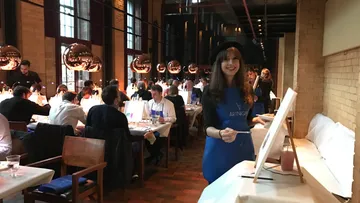 Woman painting in restaurant setting, diners in background.