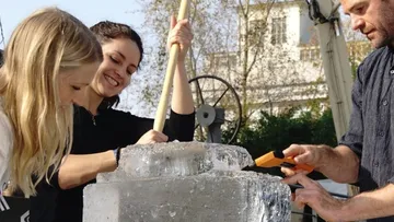 Drei Personen schnitzen draußen einen großen Eisblock.