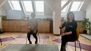 Zwei Frauen sitzen auf Stühlen und lächeln in einem Yogastudio.