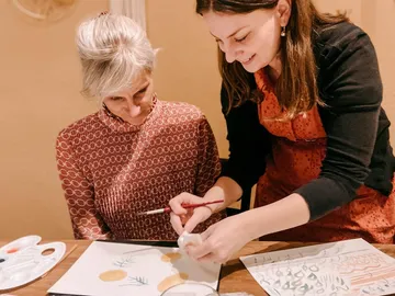 Two women paint together at a table using paints.