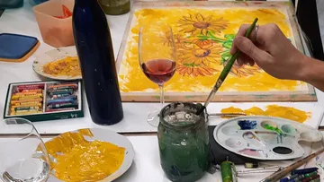 Hand painting sunflowers, wine glass nearby.