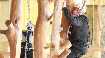 Mann mit Helm klettert auf eine Holzkonstruktion im Innenbereich.