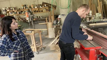 Man sanding wood, woman observing in workshop.