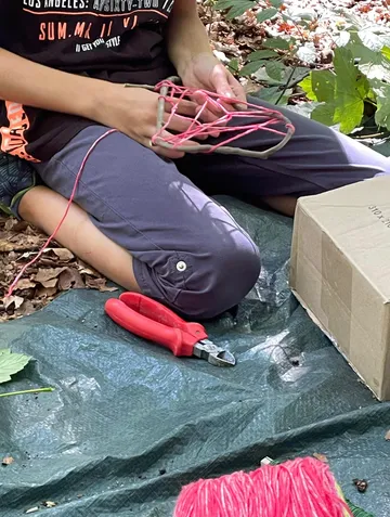 Person, die im Freien eine rosa Schnur auf Stöcken webt.