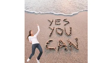 Woman on beach with 'Yes You Can' in sand.