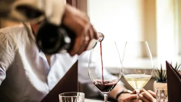 Person, die am Tisch Rotwein in ein Glas gießt.