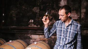 Man examining drink in cellar with barrels.