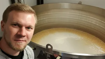 Man smiling near large fermentation tank indoors.