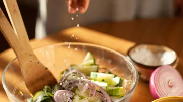 Person seasoning vegetable salad in bowl.