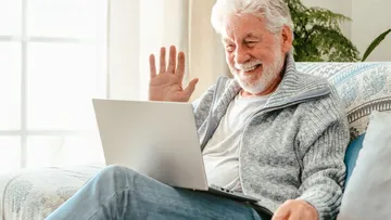Elderly man waving at laptop, sitting indoors.