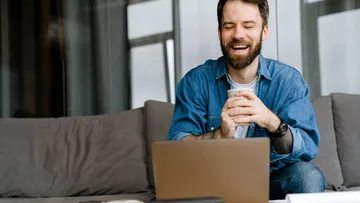 Lächelnder Mann, hält eine Tasse in der Hand und benutzt Laptop auf der Couch.