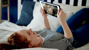 Person lying on bed using tablet for video call.