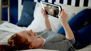 Person, die auf dem Bett liegt, ein Tablet in der Hand hält und Video schaut.