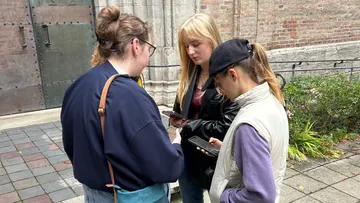 Three people looking at phones outdoors by brick wall.