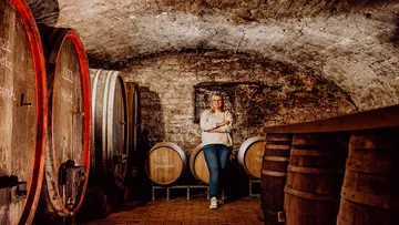 Person standing among wine barrels in a cellar.