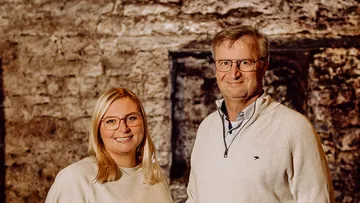 Two people wearing glasses, stone wall background.