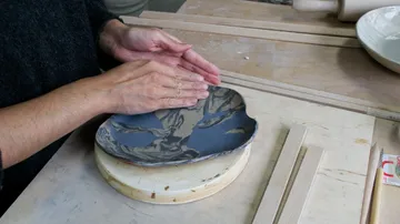 Hands shaping clay plate on wooden table.