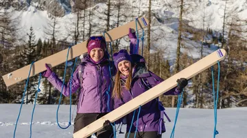 Zwei Frauen in lila Jacken mit Holzskiern in verschneiten Bergen.
