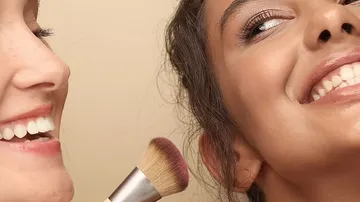 Zwei lächelnde Frauen, eine hält einen Make-up-Pinsel.