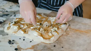 Hands prepare flatbread with toppings on counter.