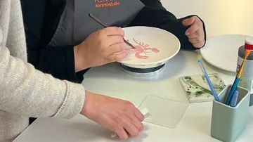 Person, die Hummer-Design auf Keramikplatte malt.