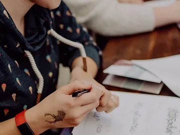 A hand with pen writes on paper at the table.