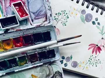 Watercolors and brushes lie next to a sketched flower illustration.