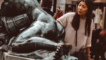 Woman gesturing at bronze sculpture outdoors.