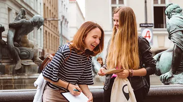 Two women laughing near a fountain in a city.